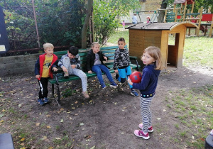 Cecylia trzyma piłkę w dłoniach stojąc na przeciwko kolegów z grupy, którzy siedzą na ławce